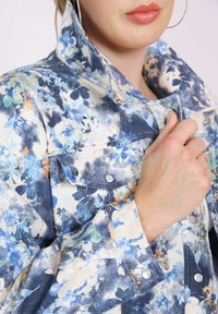 Flower-patterned denim jacket with a light blue and cream color palette. Features snap buttons and a pointed collar.