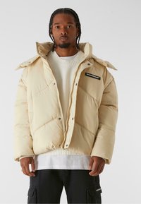 Beige padded jacket with a high collar, zip closure, and diagonal stitching. Features a logo patch on the left chest and a zippered hand pocket.