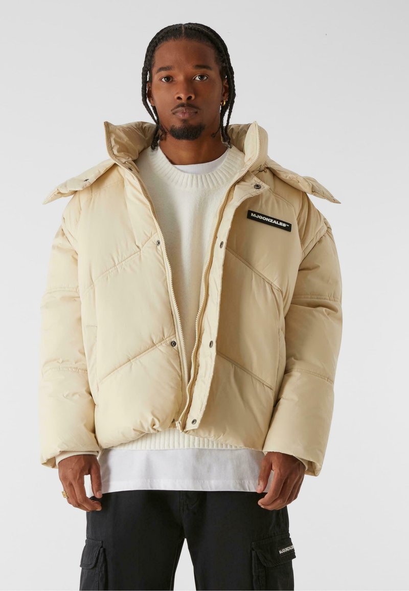 Beige padded jacket with a high collar, zip closure, and diagonal stitching. Features a logo patch on the left chest and a zippered hand pocket.