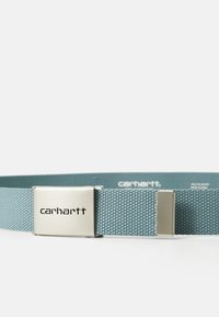 Curea din material țesut de culoare albastră, cu o cataramă metalică argintie gravată cu "carhartt". Prezintă un model texturat și o construcție robustă.