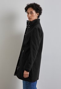 Svart kappa med hög krage, dragkedja och tryckknappar, två sidofickor och ribbade mudd. Slät textur och skräddarsydd design.