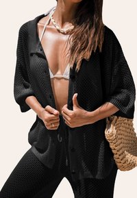 Cardigan in rete nera con una vestibilità rilassata, maniche corte e chiusura con bottoni, abbinato a un top bikini bianco e a una borsa a rete.