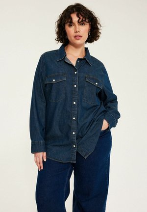 Vrouw met krullend haar die een donkerblauw denim overhemd met knopen draagt en bijpassende wijde jeans, poseert met één hand in haar zak.