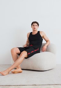 Kurkasta, off-white oblazinjena stolica; posameznik v črnem topu s tankimi naramnicami in kratkih hlačah. Stolica ima teksturirano, mehko površino; minimalistična zasnova.