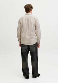 Homme aux cheveux châtain clair portant une chemise beige à rayures, un jean noir ample et des chaussures noires, debout de dos sur un fond blanc uni.
