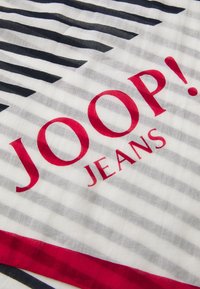 Svītrota auduma šalle ar maiņojošām melnām un pelēkām līnijām, ar izteiksmīgu sarkanu "JOOP! JEANS" logo centrā un sarkanu maliņu.
