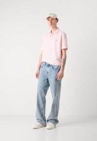 Polo shirt rose clair à manches courtes et logo discret, associé à un jean en denim bleu clair, ample et à des baskets blanches.