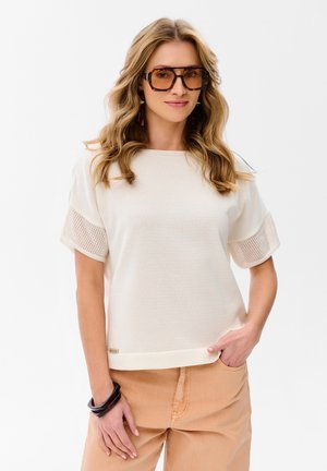 Kobieta o falujących blond włosach, nosząca duże okulary w oprawkach w kolorze żółwia, biały krótki top z detalami z siateczki, beżowe spodnie i czarne bransoletki.