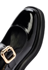 Chaussure en cuir verni noir avec un bout rond, une bride ajustable et une boucle dorée. Dispose d'une semelle noire texturée et d'une doublure intérieure douce.