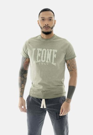Man met kort haar en baard, gekleed in een olijfgroen "LEONE 1947" T-shirt en donkere broek, met tatoeages op beide armen, kijkt recht vooruit.