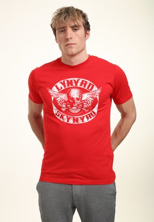 Jonge man draagt een rood Lynyrd Skynyrd T-shirt met een schedel en vleugels-graphic, gecombineerd met een grijze broek, staat met de handen achter de rug.