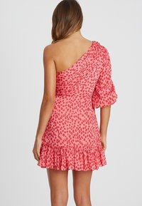 Jednobodový šaty z růžové látky s červenými abstraktními vzory, s nařaseným lemem a balónovou rukávem na jedné paži.