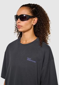 Czarne okulary przeciwsłoneczne o sportowym wzornictwie, ciemne soczewki i teksturowana czarna rama. Model nosi luźną szarą koszulkę z niebieskim haftem.