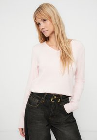 Camisola de gola em V cor rosa claro, feita de material macio, combinada com jeans de lavagem escura de cintura alta e um cinto preto com fivela dourada.