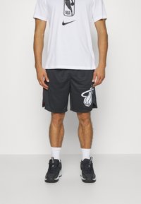 Fekete kosárlabda short, amelyen a Miami Heat logó látható, könnyű anyagból készült, sima textúrával. Fehér NBA logós pólóval párosítva.