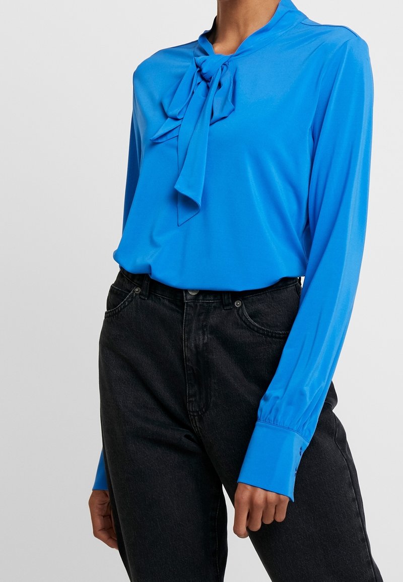 Blå långärmad blus med knytdetalj vid kragen, tillverkad av lätt tyg. Bärs med högmidjade svarta jeans.