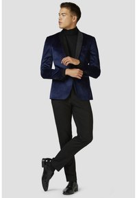 Blazer di velluto blu navy con rever in satin nero, abbinato a un dolcevita nero e pantaloni neri eleganti, completato da scarpe lucide nere.
