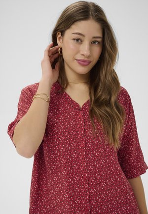 Jeune femme aux longs cheveux bruns portant un chemisier à boutons rouge à motifs, des boucles d'oreilles créoles dorées, des bracelets et un collier délicat.