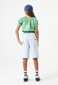 Blusa verde corta con puños elásticos, pantalones cortos de mezclilla azul claro, zapatillas negras y calcetines blancos con logo. Vista trasera del atuendo.