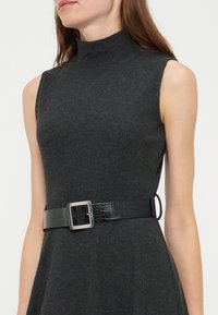 Femme portant une robe sans manches en maille côtelée gris foncé avec un col haut et une ceinture noire texturée avec une boucle carrée argentée.