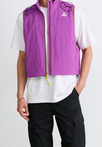 Fioletowa lekka kamizelka z zamkiem z przodu, siateczkowymi wstawkami na ramionach i żółtymi sznurkami, noszona na białej koszuli.