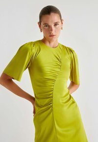 Abito verde lime con maniche a sbuffo corte, caratterizzato da arricciature laterali e una texture liscia, creando una silhouette aderente.