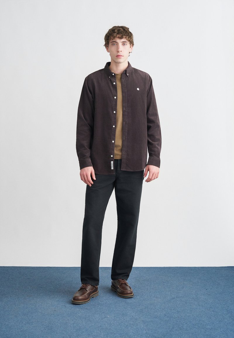 Camicia marrone scuro con bottoni, in tessuto strutturato, indossata sopra una camicia color sabbia. Abbinata a pantaloni neri e scarpe marroni con lacci.