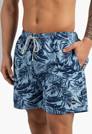 Homme portant un short de bain bleu à imprimé tropical avec cordon, main dans la poche et bras tatoué visible.