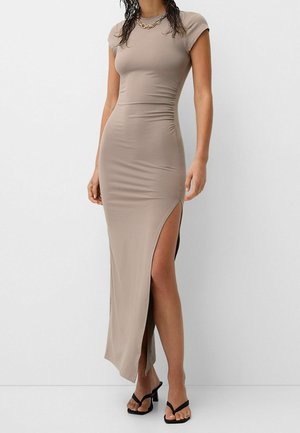 Femme portant une robe maxi taupe ajustée à manches courtes avec une fente haute sur le côté et des sandales à talons noires sur un fond blanc uni.