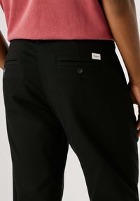 Zwarte katoenen broek met een gladde textuur, voorzien van twee achterzakken en een klein merklabel boven de rechterzak.