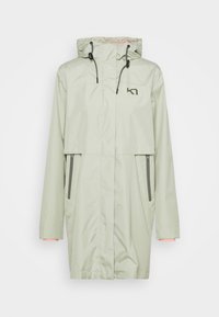 Ljusgrön regnjacka med huva, två sidofickor med dragkedja och kontrasterande innerkant på ärmarna. Inkluderar en logotyp på bröstet. Slätt, vattenavvisande material.