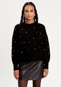 Pull en maille noir avec un col rond, agrémenté de décorations argentées éparpillées, associé à une jupe courte en cuir noir.