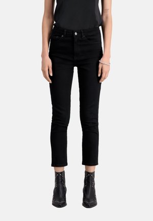 Slim-fit black jeans worn with black studded ankle boots and a black sleeveless top, showing arms and midsection.