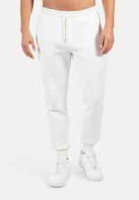 Pantalones deportivos blancos hechos de tela suave con cintura elástica y cordón, con piernas entalladas y puños acanalados. Combinados con zapatillas deportivas blancas.