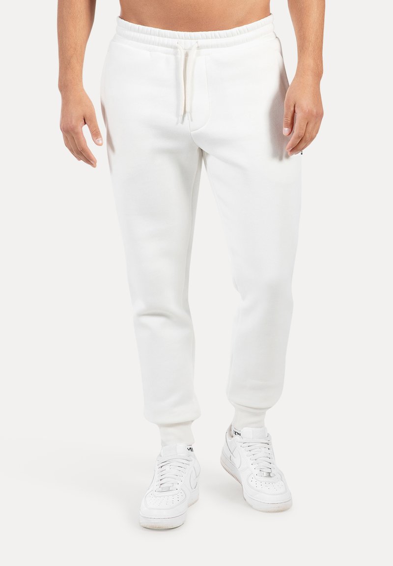 Pantalones deportivos blancos hechos de tela suave con cintura elástica y cordón, con piernas entalladas y puños acanalados. Combinados con zapatillas deportivas blancas.