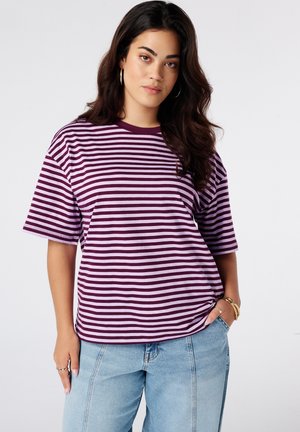 Jonge vrouw met lang donker haar die een paarse en lichtroze gestreepte T-shirt en lichtblauwe spijkerbroek draagt, staand met één hand in de zak.