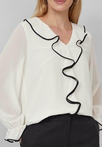 Weiße Bluse mit schwarzem Rüschenbesatz entlang des Ausschnitts und der Ärmel. Durchsichtiges, leichtes Material mit gerafften Ärmelbündchen. Kombiniert mit dunklen Hosen.