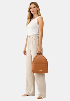 Woman wearing white sleeveless top and beige wide-leg pants, holding a brown leather backpack with gold logo by its handle.