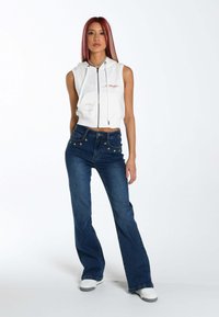 Sudadera sin mangas de color blanco y corte corto con cremallera frontal y detalles bordados, combinada con unos vaqueros acampanados de color azul oscuro con acentos de estrellas.