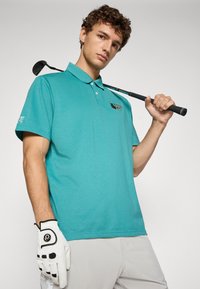 Turkis polo skjorte med krage, broderte detaljer på brystet, iført hvite golfhansker, som holder en golfklubbe over skulderen.