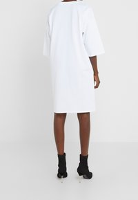 Weißes oversized T-Shirt-Kleid mit kurzen Ärmeln, rundem Ausschnitt und gerader Silhouette. Trägt dazu schwarze Knöchelschuhe.