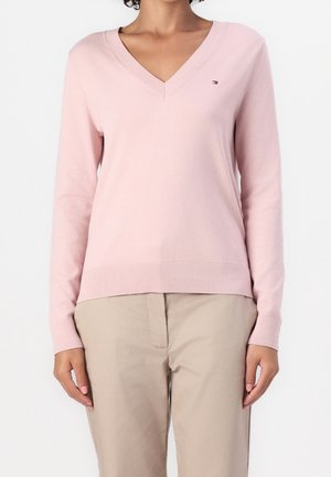 Personne portant un pull rose clair à col en V et manches longues avec un pantalon beige, debout devant un fond uni et clair.