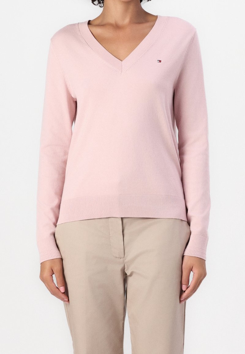 Personne portant un pull rose clair à col en V et manches longues avec un pantalon beige, debout devant un fond uni et clair.