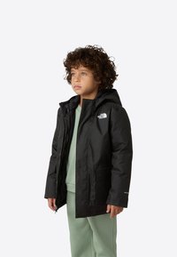 Černá voděodolná bunda s kapucí, předním zipem a dvěma kapsami. Nositá přes světle zelenou mikinu a odpovídající kalhoty.