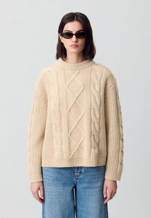 Un pull en tricot beige avec une texture épaisse, présentant un motif en torsades et un col rond. Associé à un jean large bleu clair.