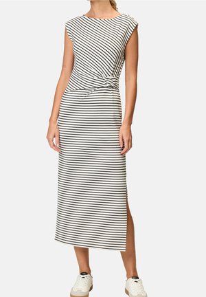 Kobieta ubrana w czarno-białą, poziomo paskowaną sukienkę midi bez rękawów z bocznym rozcięciem i białe trampki, stojąca na tle jednolitego tła.