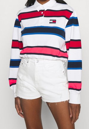 Kobieta ubrana w biały polo z długim rękawem ozdobione czerwonymi, niebieskimi i czarnymi paskami, w połączeniu z białymi, wysokim stanem szortami jeansowymi ze strzępionymi brzegami.