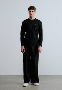 Musta pitkähihainen paita ja leveälahkeiset housut, molemmat sileää kangasta. Paidassa on pieni logo rinnassa. Valkoiset kengät.