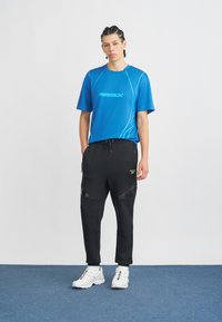 T-shirt atlético azul com logótipo e linhas de contraste, combinado com calças de jogging pretas com bolsos com fecho e acabamentos brilhantes. Ténis brancos.