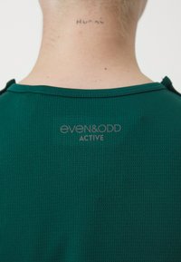 Zelena aktivna majica izrađena od teksturirane tkanine. Sadrži "even&odd ACTIVE" logo na vratu i mali detalj dugmeta na ramenu.